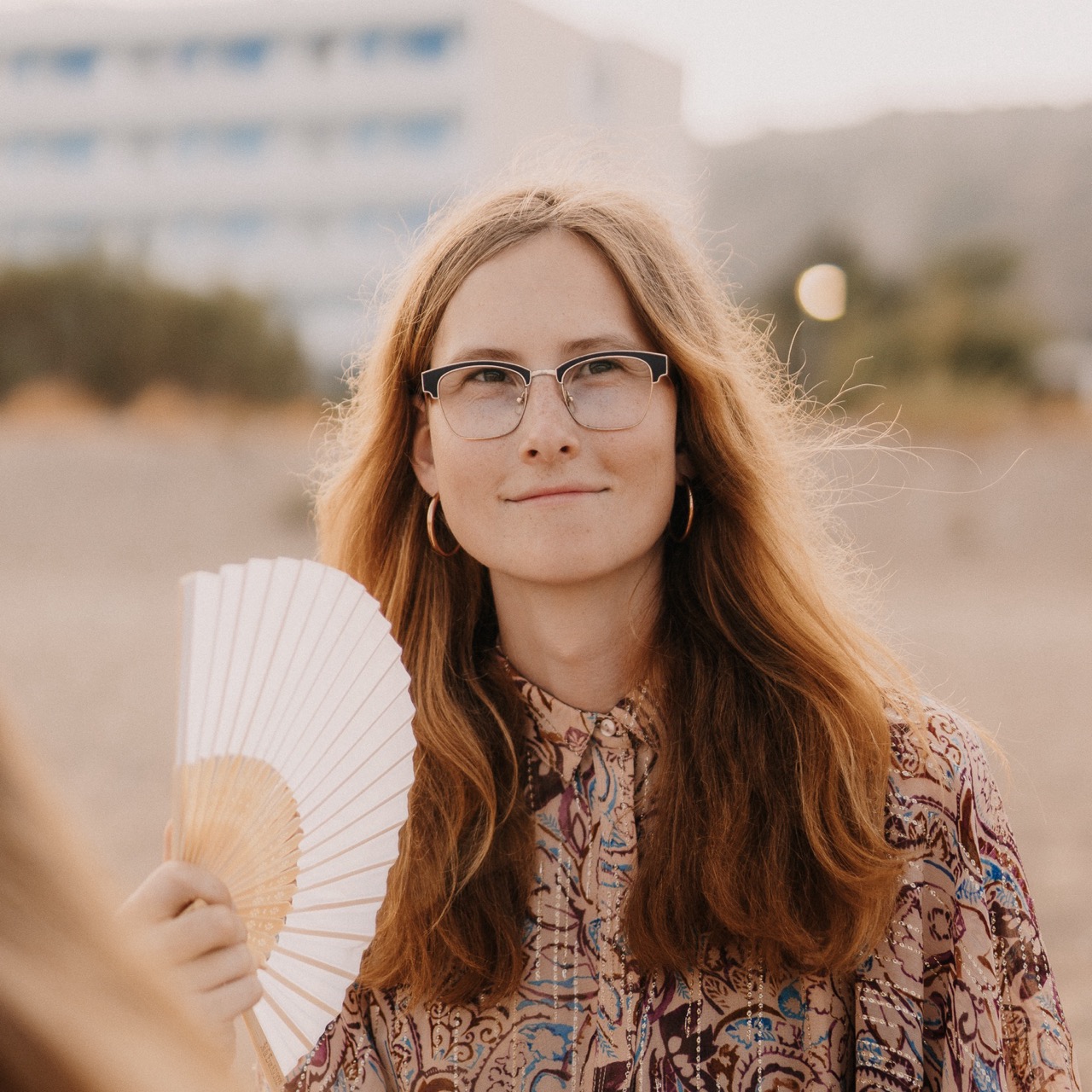 A photo of Jocy sitting on a beach in a beige dress. Looking next to the camera. Holding a hand fan in her hand